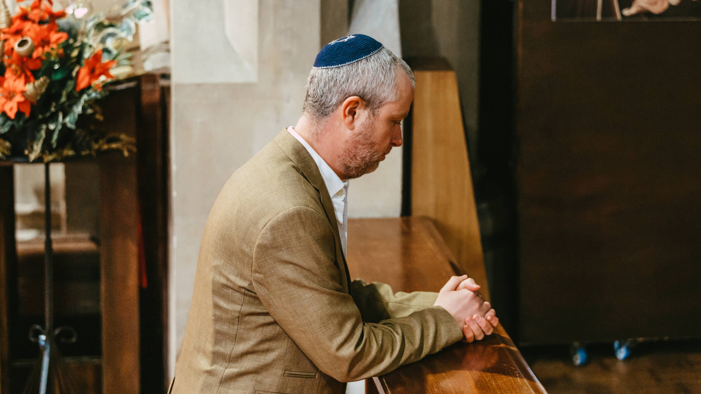 Portrait of a Jewish man kneeling and praying in the synagogue. The man is wearing a yarmulke on his head as he sits alone in the synagogue.