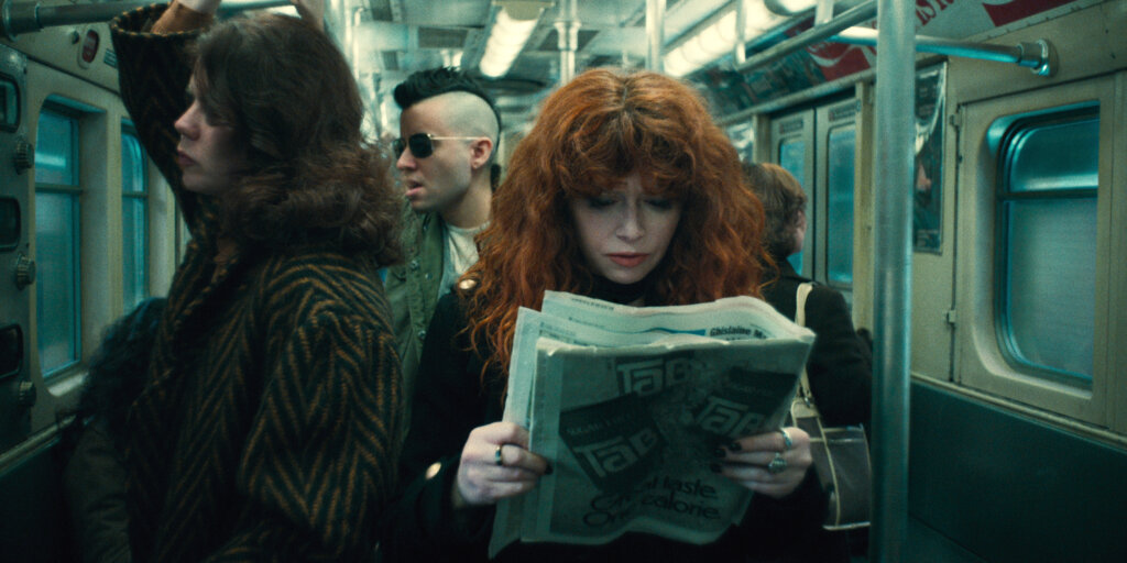 Natasha Lyonne, with redheaded curls, stands on an old subway car looking down at a newspaper. A man with a mohawk stands behind her.