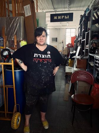 Woman wearing t-shirt with hebrew letters on it stands smiling with hands on hips. In background, a long room with art supplies.