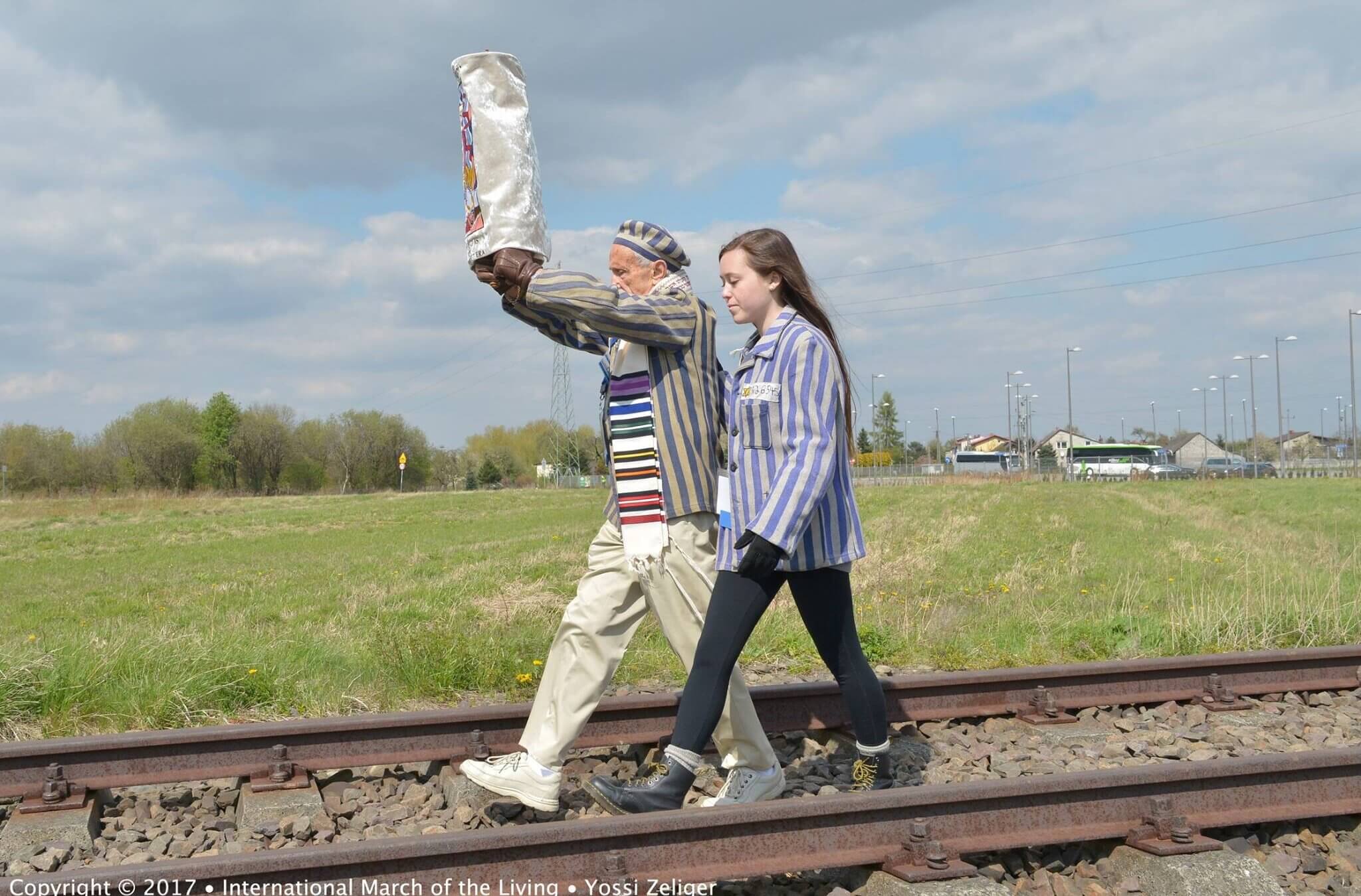 A Holocaust survivor holds a Torah scroll aloft as he walks with a young woman down the train tracks leading from Auschwitz to Birkenau.