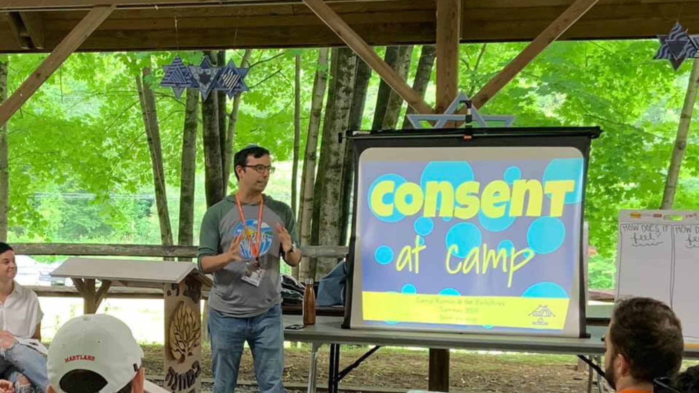 Rabbi Ethan Linden, director of Camp Ramah in the Berkshires, gives a presentation at an outdoor venue standing in front a screen that says "Consent at Camp."