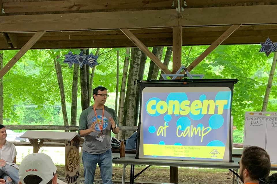 Rabbi Ethan Linden, director of Camp Ramah in the Berkshires, gives a presentation at an outdoor venue standing in front a screen that says "Consent at Camp."