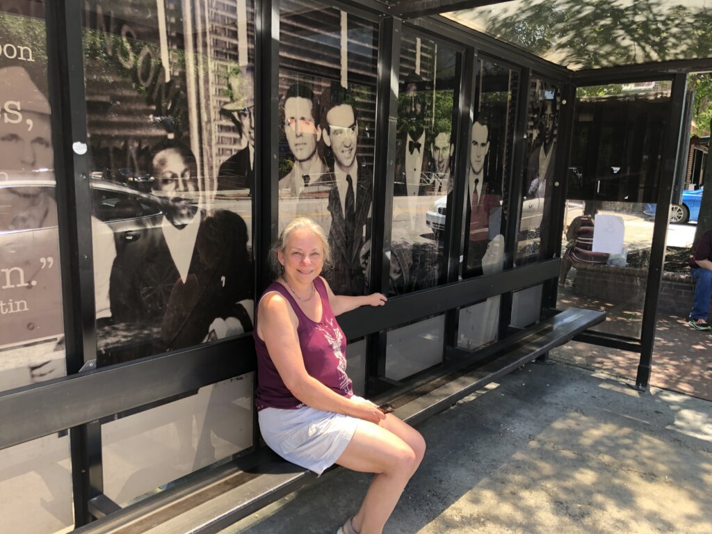 Amy Zowniriw at the bus stop where her uncle, Igor Roodenko, was arrested. Her uncle and others from that Freedom Ride are on the poster behind her.