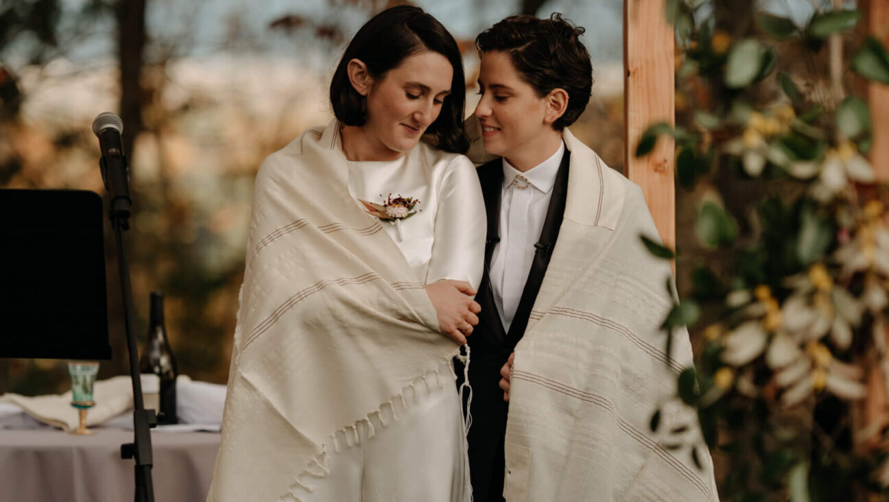 Two women stand together, one in a long white gown and the other in a dark suit, wrapped in a tallit.