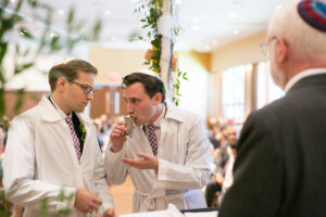 Two men in white robes, one drinking from a kiddush cup before