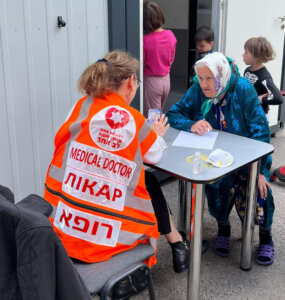 Woman in orange safety vest seated at a table, typing on a cellphone, facing an old woman with a headscarf