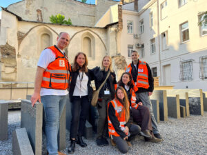 Six people, some in orange safety vests, pose in front of an arcaded building.