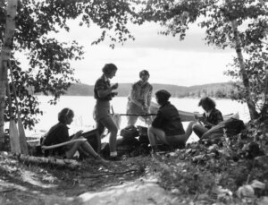 Five women campers sitting around campfire in front of lake