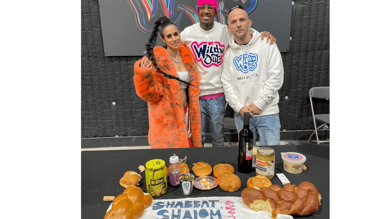 Woman in orange coat with two men stand behind a Shabbat Shalom spread