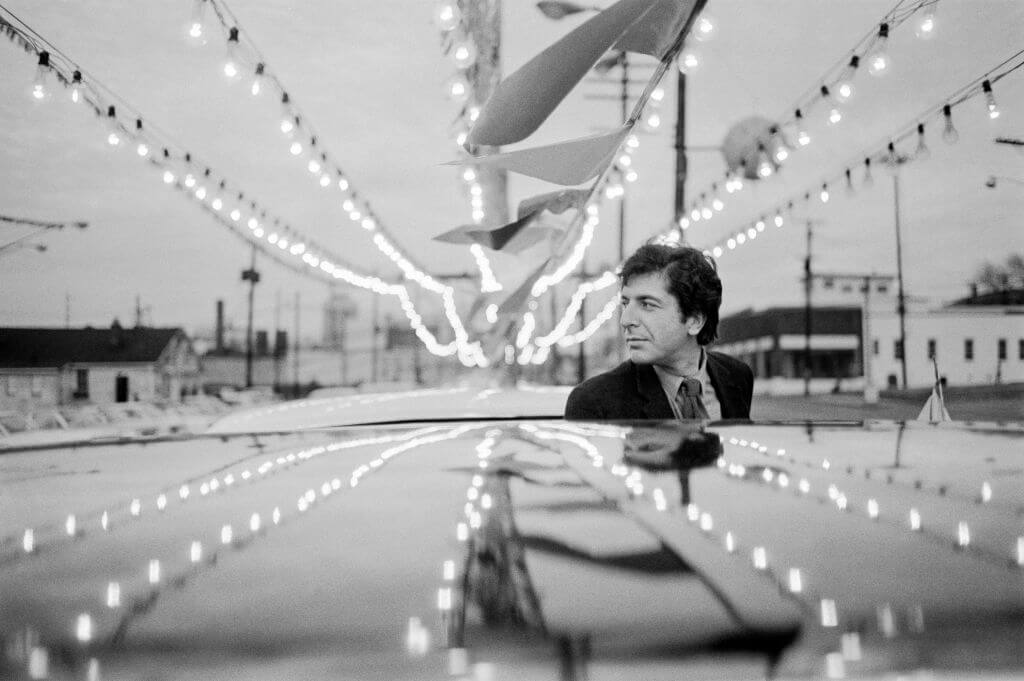 Singer-songwriter Leonard Cohen standing behind a car in a black and white photo