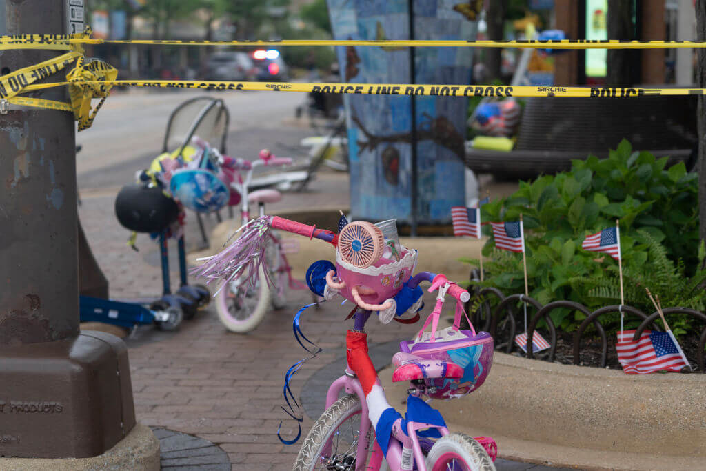 The picture shows a child's bicycle, left behind on the sidewalk after the mass shooting at Highland Park, next to a strip of "caution" tape.