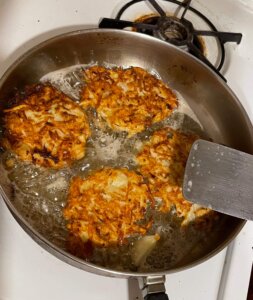 Frying latkes in a skillet, puffs of flour and oil cobwebbing out onto the  stovetop. The cook flipping the potato pancakes with a spatula, kids  eagerly peeking inside the skillet. Generative AI