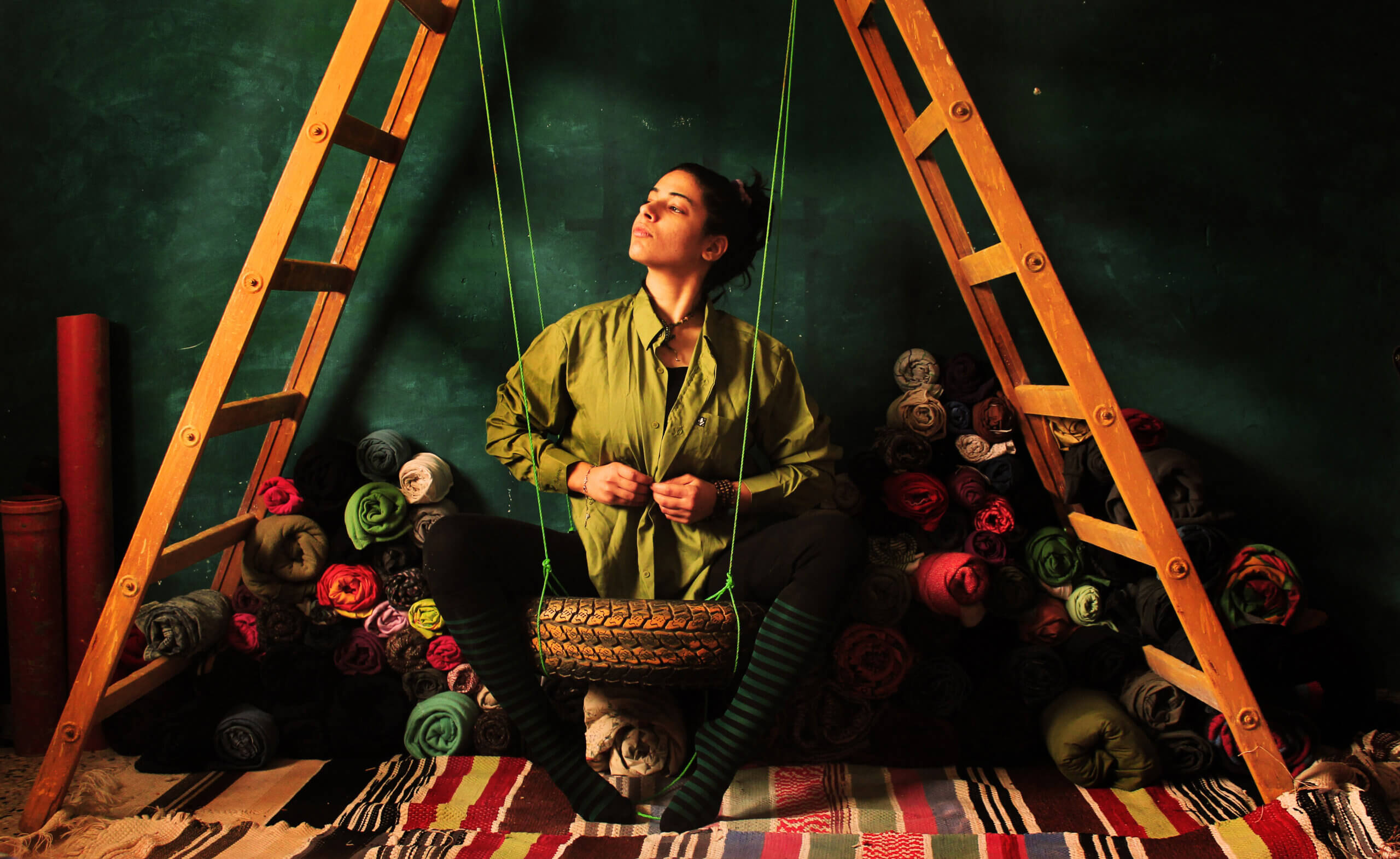 A self-portrait of artist Nidaa Badwan, underneath a ladder, in front of a miniature tire swing, surrounded by mounds of fabric