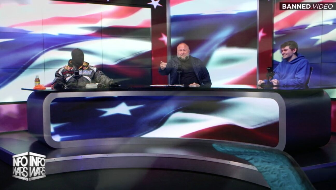 Kanye West, in a full-face mask, seated at the Infowars table with Alex Jones, in middle, and Nick Fuentes, at right. A projection of the American flag is on a screen behind them.
