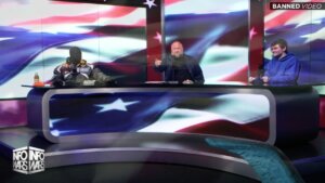 Kanye West, in a full-face mask, seated at the Infowars table with Alex Jones, in middle, and Nick Fuentes, at right. A projection of the American flag is on a screen behind them.