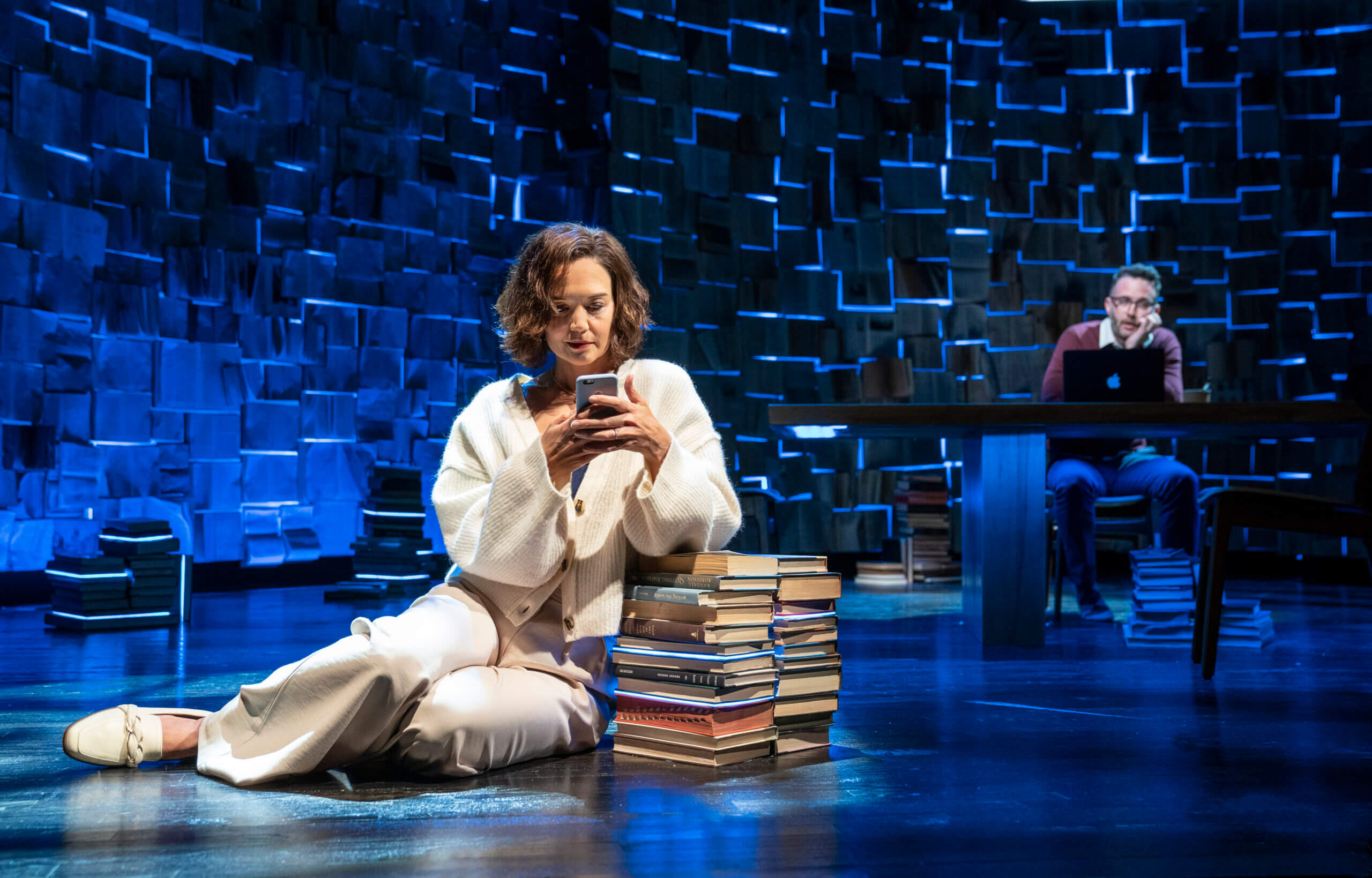 A female actor in a white cardigan and pants sits on the floor near a pile of books, looking at an email on her phone. 