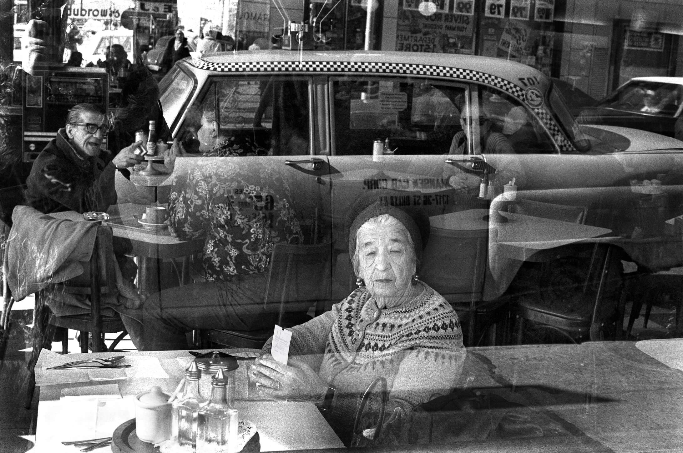 An elderly woman sitting at a table is photographed through a window, with the reflection of the street outside superimposed on her face.