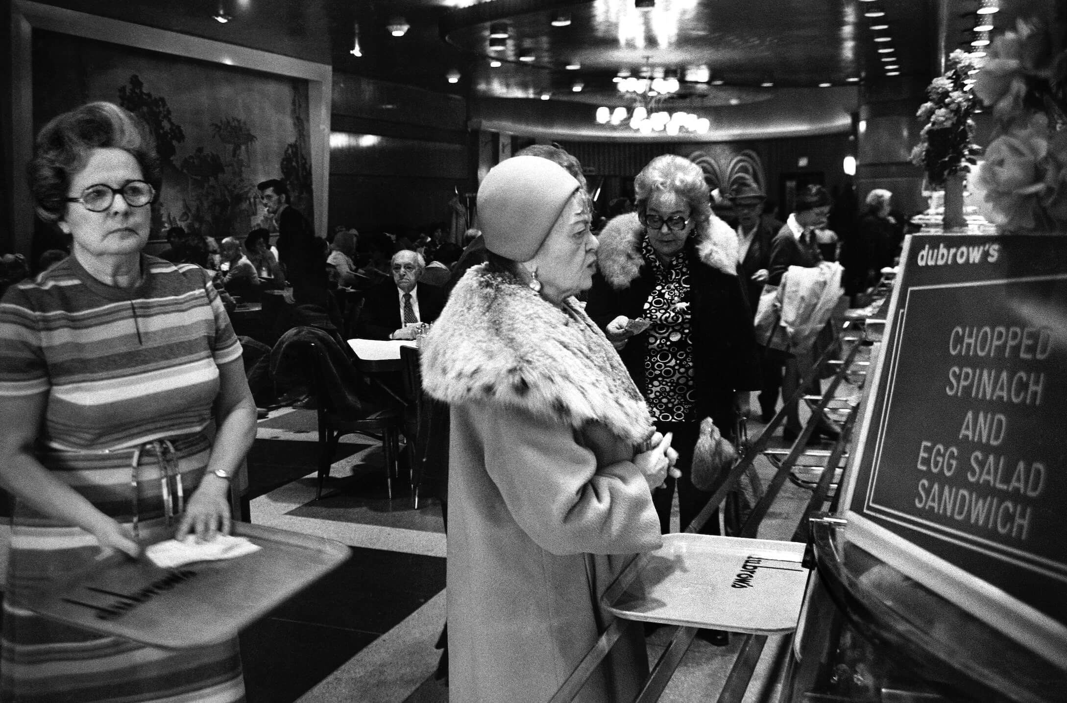 A woman dressed in a formal coat and cloche hat examines the menu at Dubrow's Cafeteria.