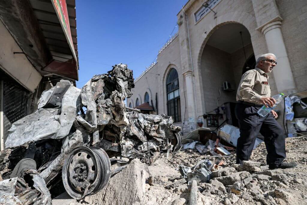 People walk past rubble and the remains of a destroyed vehicle outside a mosque in the occupied West Bank city of Jenin on July 5, 2023, after the Israeli army declared the end of a two-day military operation in the area. 
