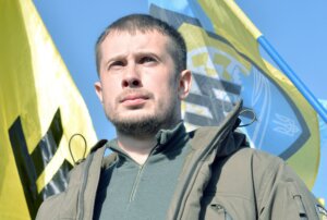 Andriy Biletskiy in an olive green jacket, staring with an upward gaze in front of Azov battalion flags.