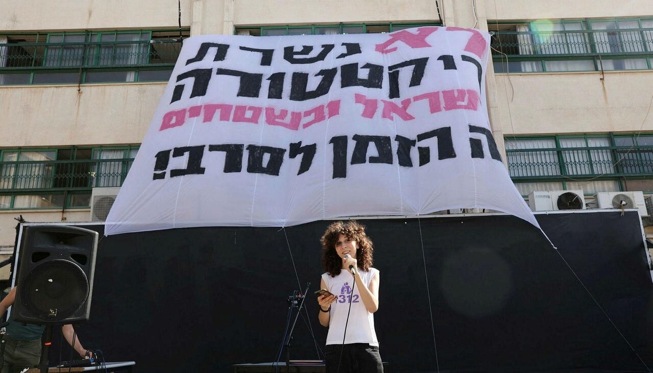 A student standing in front of a banner that says 'We won't serve the dictatorship in the West Bank and Israel, it's time to refuse,' on Sep. 3, 2023.