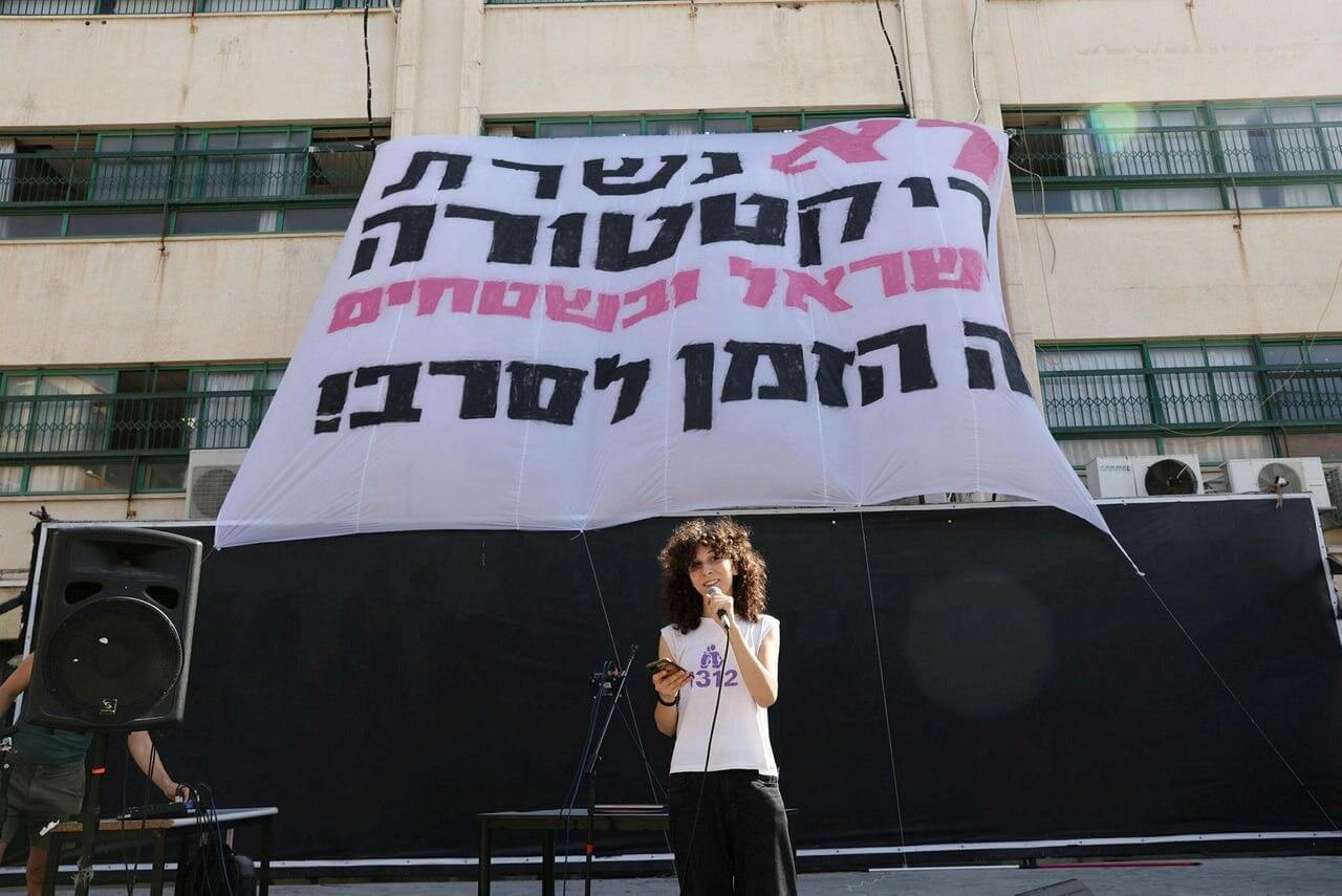 A student standing in front of a banner that says 'We won't serve the dictatorship in the West Bank and Israel, it's time to refuse,' on Sep. 3, 2023.
