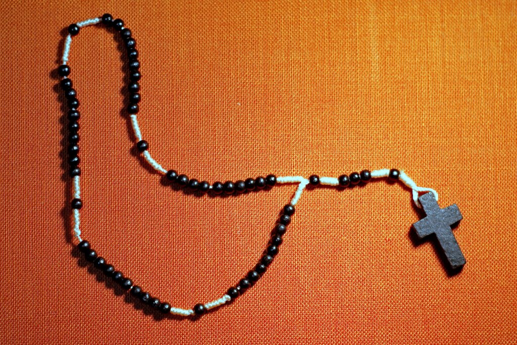 A beaded black necklace on a white string with a black, minimalist cross at the end, all on a pumpkin-colored background.