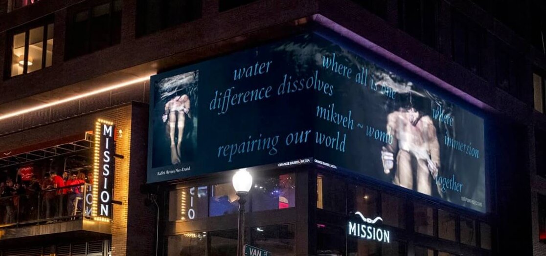 A street corner with, on the left, a restaurant with a vertical sign that reads "Mission." On the corner, a dark blue digital panel with an image of a woman curled into a ball, bathing, captioned "Rabbi Haviva Ner-David," and the words "water...where all is one...where difference dissolves...immersion...mikveh~womb...repairing our world...together" in aqua blue.