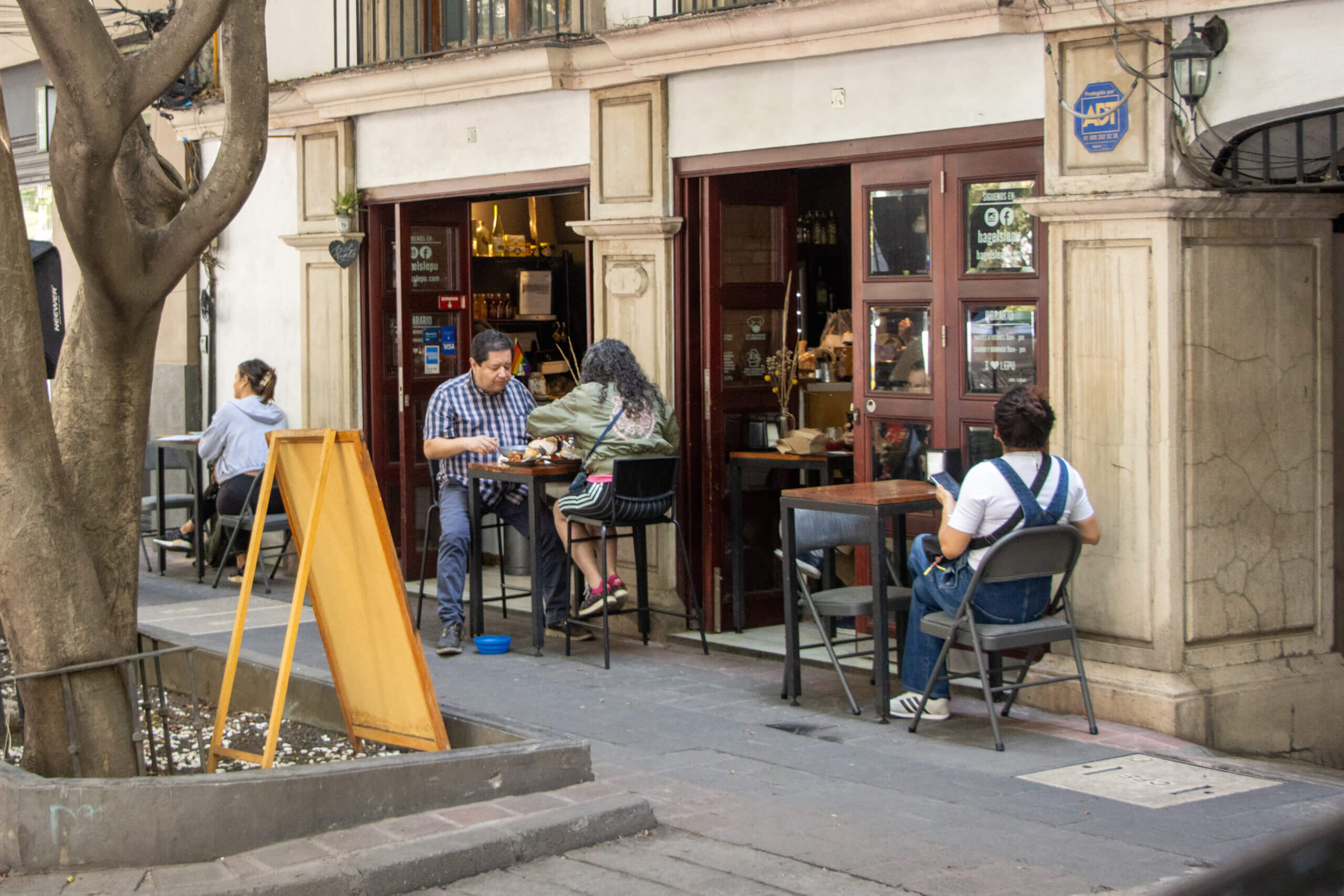 People sit at high tables on the sidewalk in front of a storefront with carved stone pillars and wooden french doors.