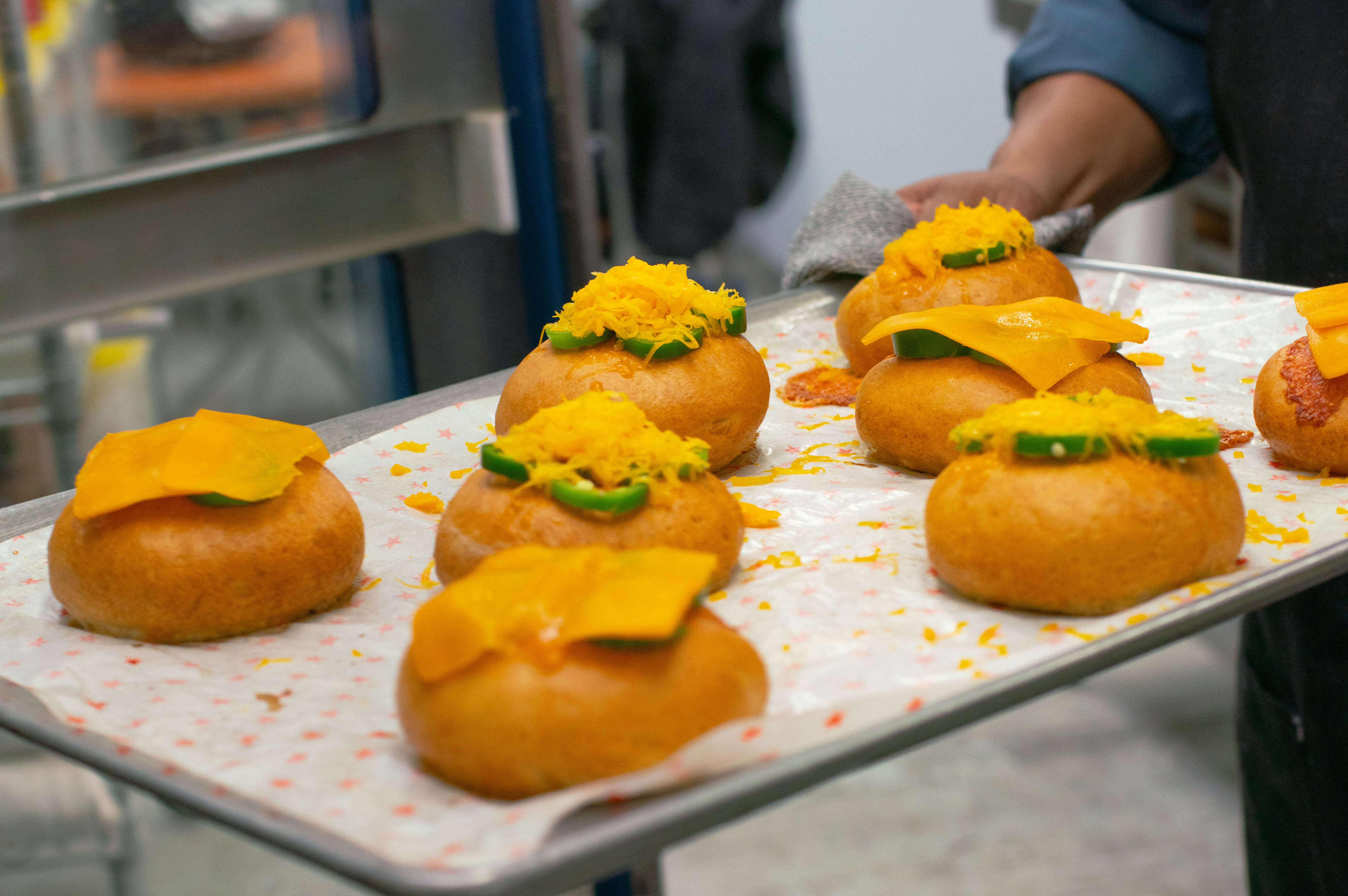A baker holds a sheetpan full of tan bagels topped with jalapeño slices and cheddar cheese.