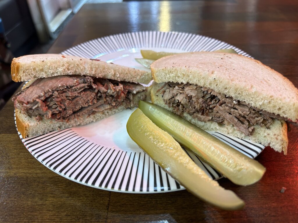 Two halves of a brisket sandwich on a white plate with radial black stripes, with two long pickles slices in front of it.