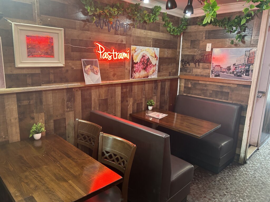 A restaurant booth with a wooden table in a corner of a restaurant with wood-paneled walls. A neon sign says "Pastrami" in handwritten print above the booth. To the left of the booth is another wooden table, with wooden chairs. Synthetic vines line the corners of the ceiling. 