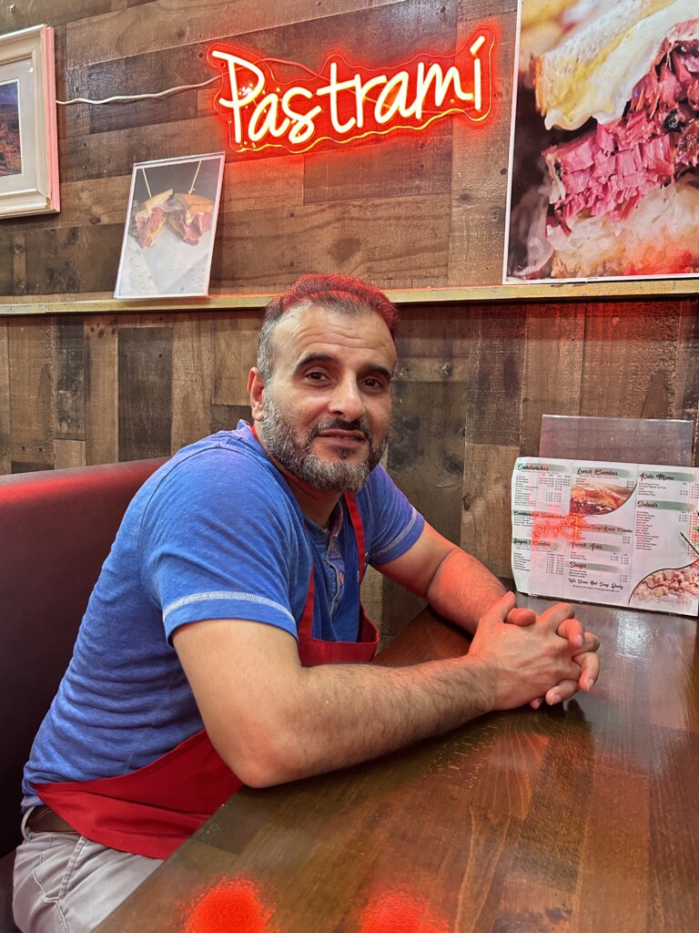 A man with light brown skin, a gray beard, thin, short, gray hair, and a blue T-shirt smiles while sitting in a restaurant booth. Above him, a red neon sign that reads "Pastrami" in handwritten print hangs on the wall, alongside photos of pastrami sandwiches.