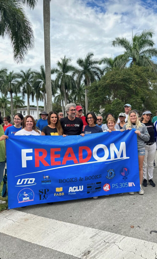 A group of people of different ages stands behind a blue banner that reads "FREADOM" on a street with rows of palm trees behind them.