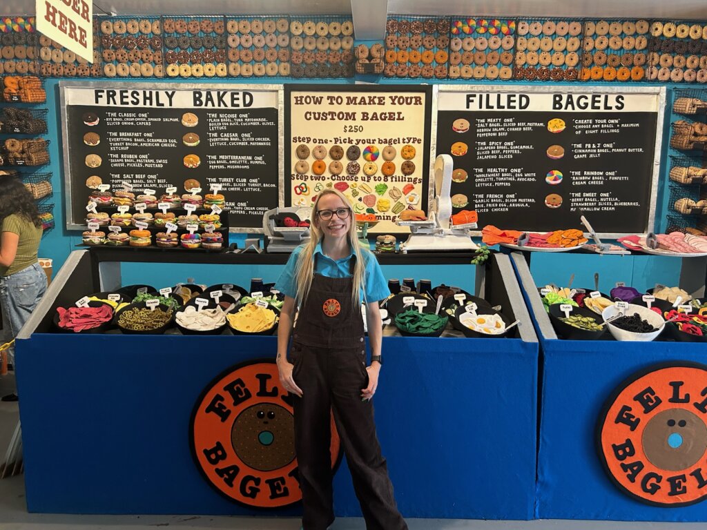 Woman in apron stands smiling in front of a deli counter with felted sandwich fillings like eggs, swiss cheese, and spinach. In the background, a sign reading "FRESHLY BAKED" and another one reading "FILLED BAGELS" with paintings of bagel sandwiches and descriptions like "The Classic One: rye bagel, cream cheese, smoked salmon, sliced onion, capers;" and "The Mediterranean One: Egg Bagel, Omelette, Hummus, Peppers, Mushrooms." Another sign reads: "How to make your custom bagel ($250): step one: pick your bagel type; step two: pick up to eight fillings."