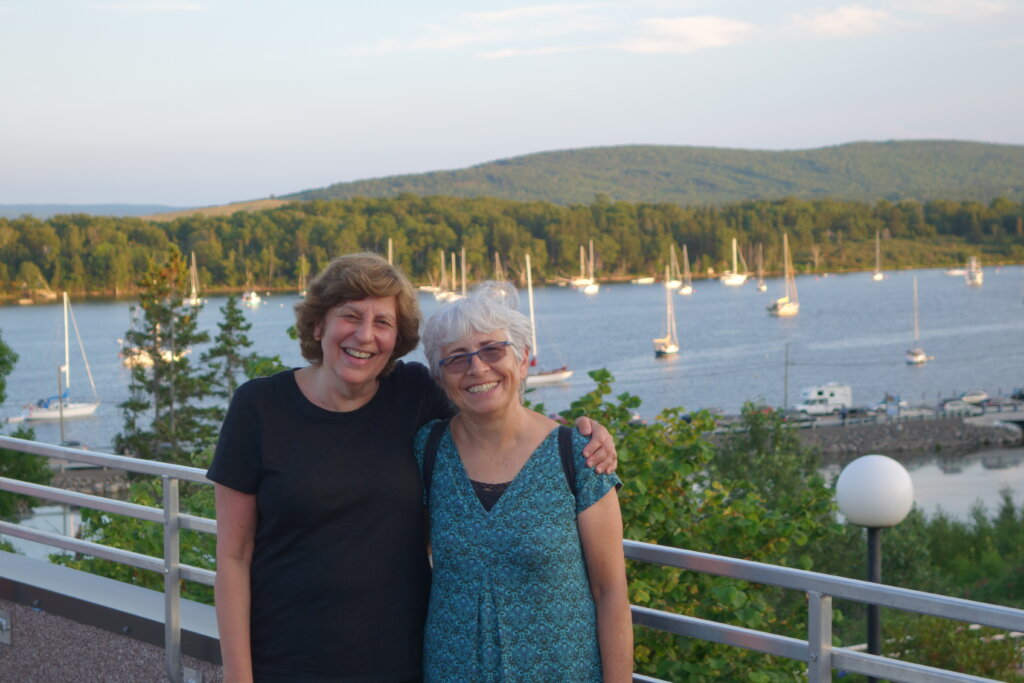 Two White women pose for a photo with their arms around each other with a white railing behind them, and a lake with sailboats ringed by trees and hills in the distance.