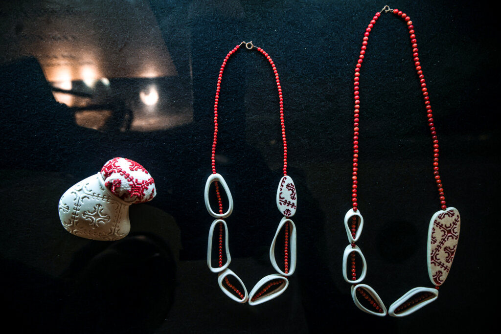 Left, a small white ceramic container with an expanding embroidered red and white cloth poking out of its opening. Center and right, two necklaces with red beaded strings, hollow white carved ceramic pendants, and white ceramic pendants etched with thin red floral patterns.