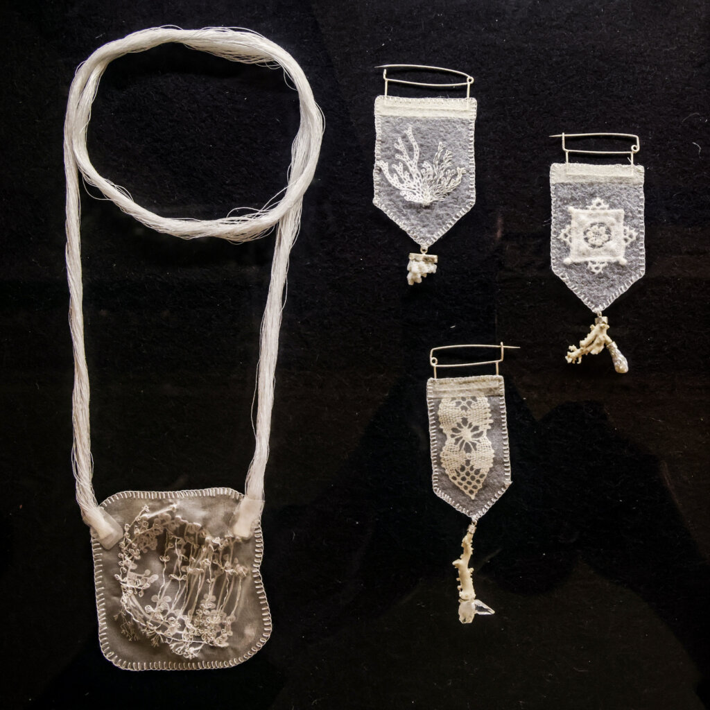 On the left, a white necklace with a thick rope made of many thin white string, and a square cloth pendant embroidered with wildflowers. On the right, three white silk pendants embroidered with coral and flowers, and affixed to little pieces of coral.
