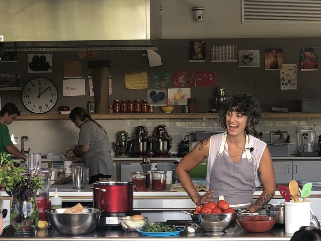 Woman smiles in apron behind kitchen island with bowls of tomatoes, greens, and other foods. Behind her, a wall full of kitchen tools like stand mixers, jars of a red sauce, and two people leaning over a sink.