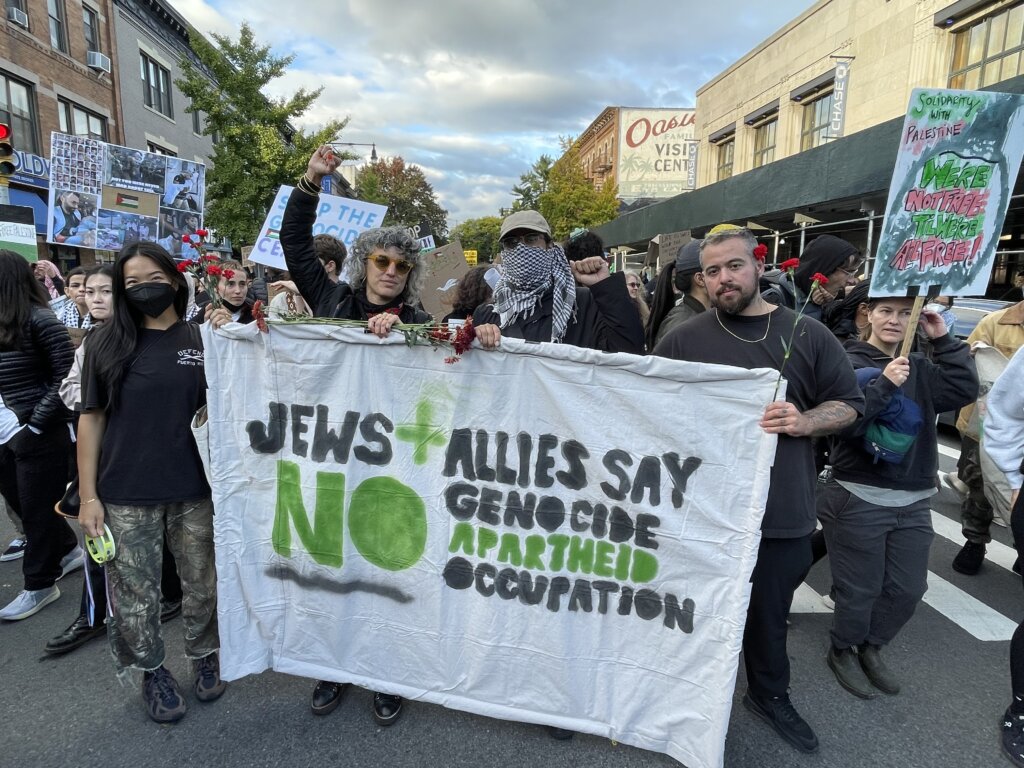 A group of people stand behind a banner that says "Jews and Allies say NO to Genocide, Apartheid, Occupation." Many people stand behind and around them, many of them holding picket signs, with buildings and trees in the background.