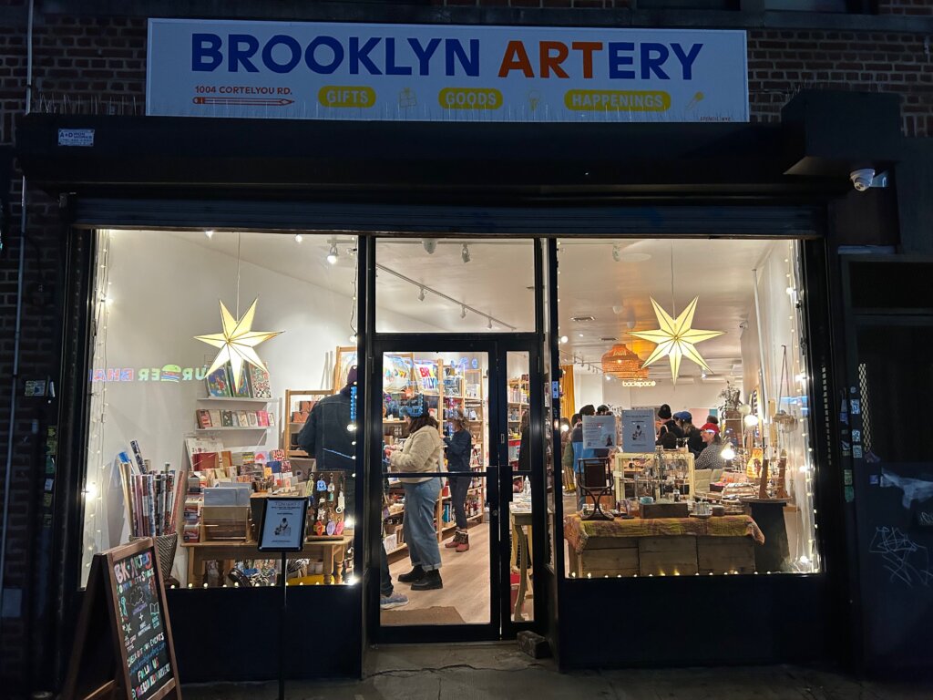 A lit-up storefront at night, full of shelves of colorful gifts and people shopping inside. In front, a sign that says "Brooklyn Artery: Gifts; Goods; Happenings."