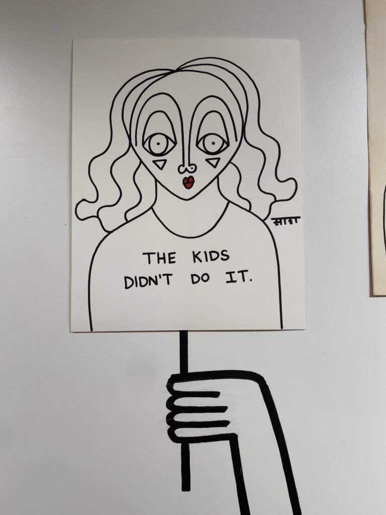 A black-and-white line drawing of a hand holding up a sign with a black-and-white line drawing of a woman's portrait on it. On her chest, the words "The kids didn't do it" are inscribed.