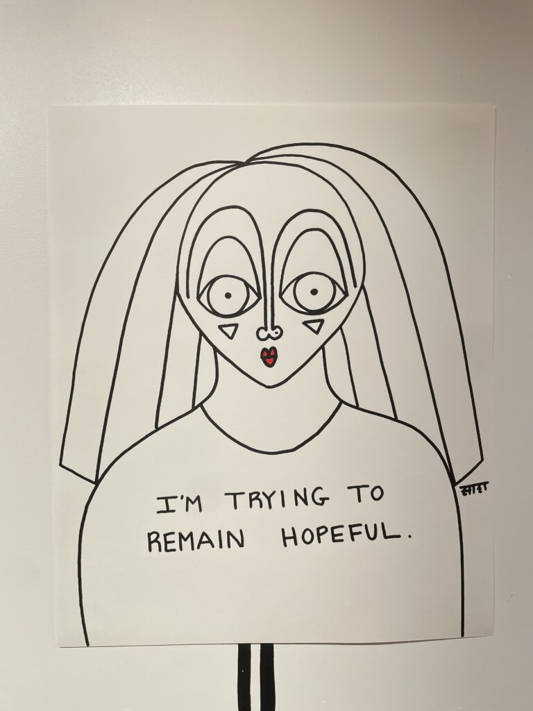 A line drawing of a woman's portrait on white paper with the words "I'm trying to remain hopeful" written on her chest.