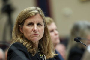 Liz Magill, a white woman in a suit, scowling while sitting in a hearing in front of a microphone.
