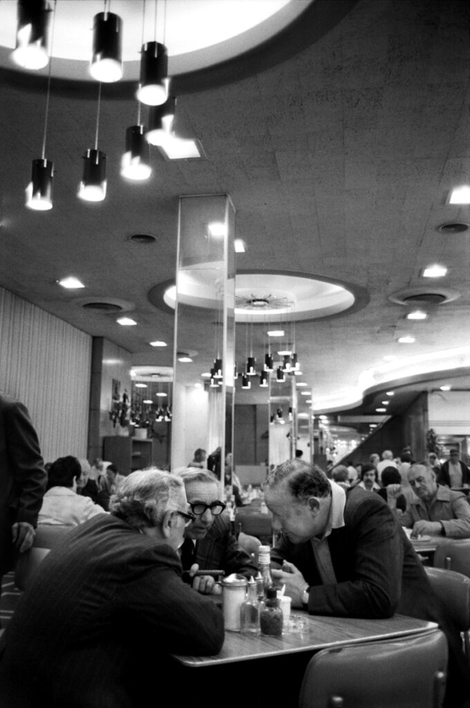 In the foreground, three middle-aged men lean over a small table to talk with their faces close to one another. In the background, modern, cylindrical chandelier fixtures, mirrored columns, and people filling other tables talking and eating.