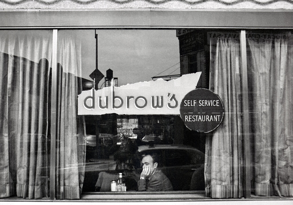 A storefront window with a decal that says "dubrow's: self-service restaurant." A man sits at a table behind the window, his left hand half covering his face. The windows are partially obscured by curtains on both sides, but open in the middle.