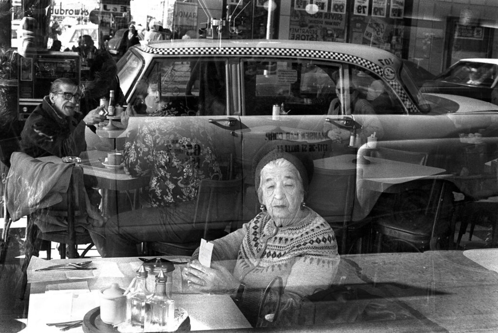 A shot through a large window of an older woman sitting at a table looking outwards with a check in her hand. Behind her, two people sit in impassioned conversation at antoher table. A reflection of a yellow taxi cab stretches over their images.