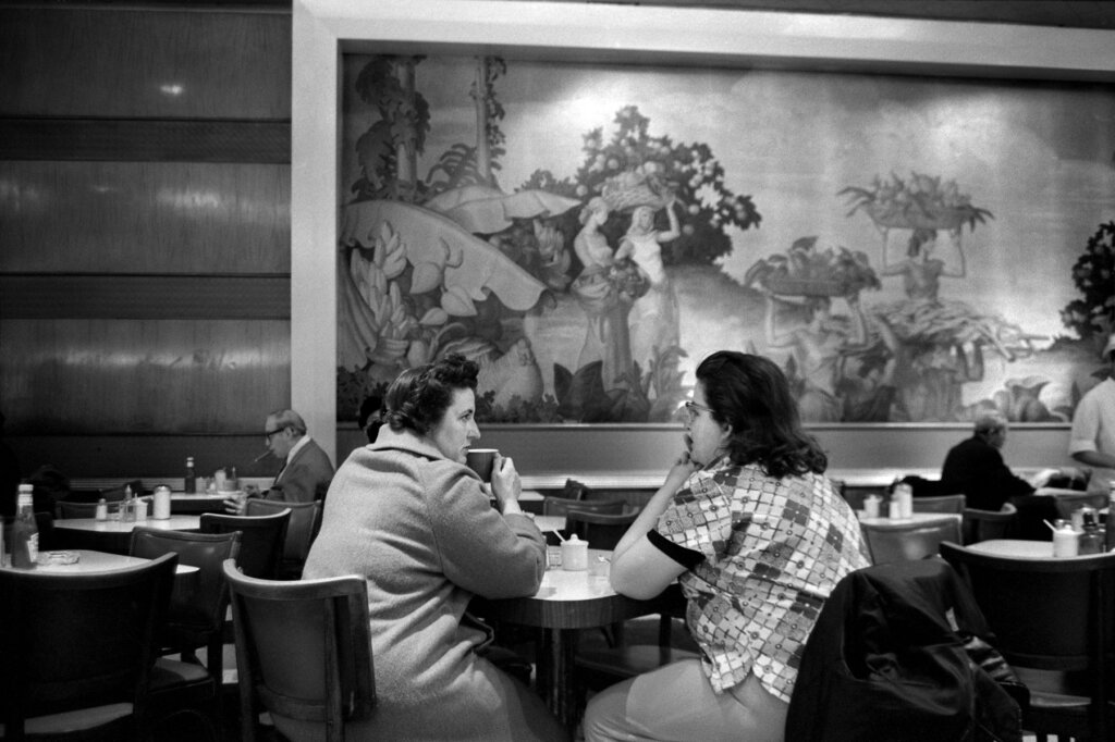 Two women sit hunched over a table with their backs turned to the camera, one of them nursing a cup of coffee. Behind them, tables and a mural of women in a seaside pastoral environment carrying baskets of fruits and vegetables.
