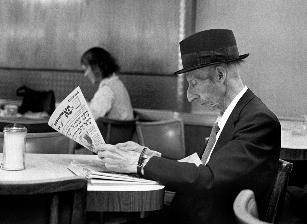 Side view of a man in a suit and top hat at a table, holding a newspaper with mostly Yiddish text and the English word "Forward" in the corner. Resting on the table is a copy of the New York Times.