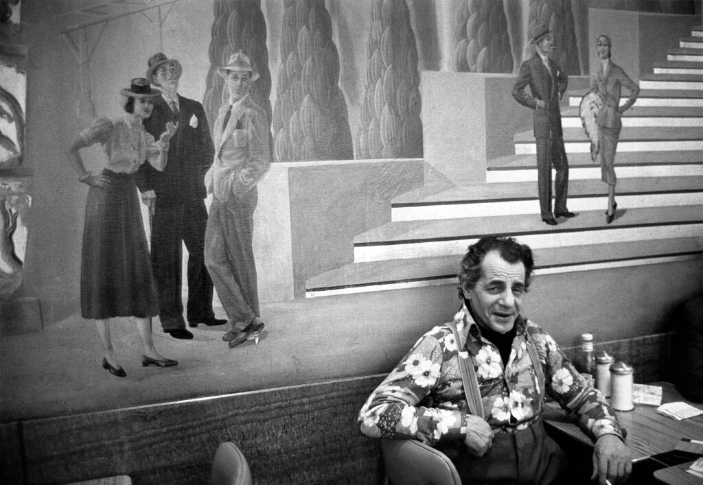 A man with suspenders and a flower shirt sits sideways on a chair, leaning against the wall and scowling. Behind him the wall is covered with a mural featuring people dressed in top hats, shirt-skirt combos, and suits around large, architectural steps.